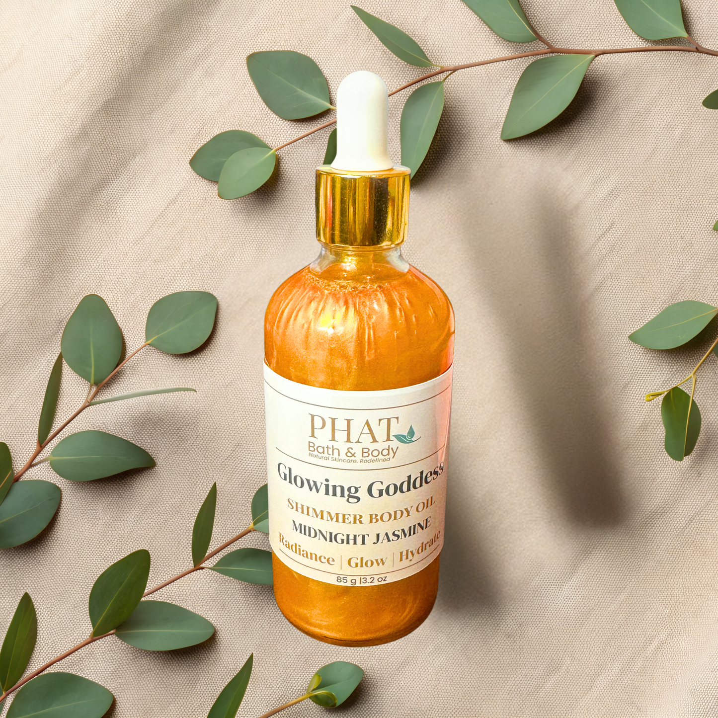 Bottle of 'Glowing Goddess' shimmer body oil from PHAT Bath & Body on a beige fabric background with green leaves.