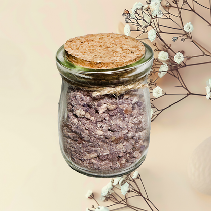 Glass jar with cork lid containing a purple bath salt mix, surrounded by white flowers on a beige background.