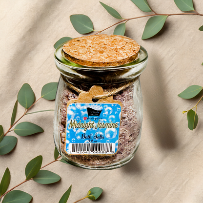 Glass jar of 'Midnight Jasmine' bath salts with a label on a beige fabric background with eucalyptus leaves.