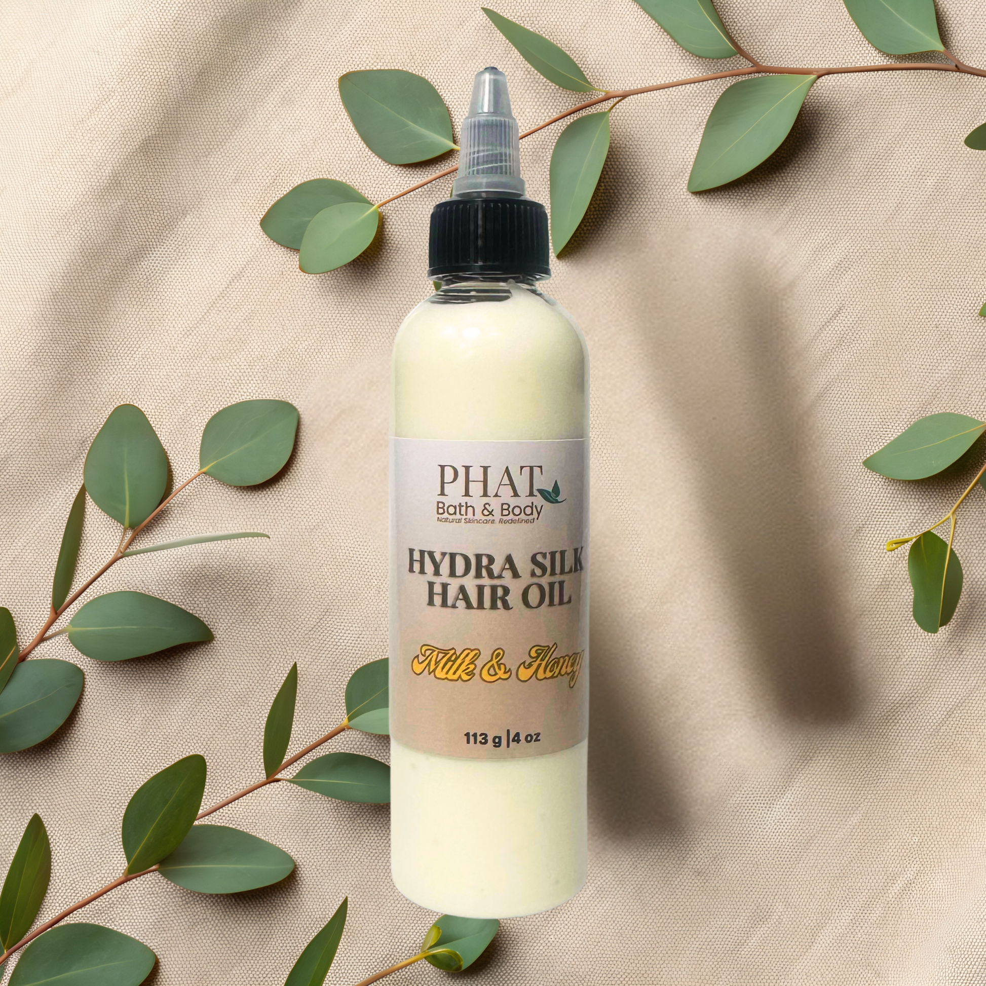 Bottle of Phat Bath & Body Hydra Silk Hair Oil with green leaves on a beige fabric background