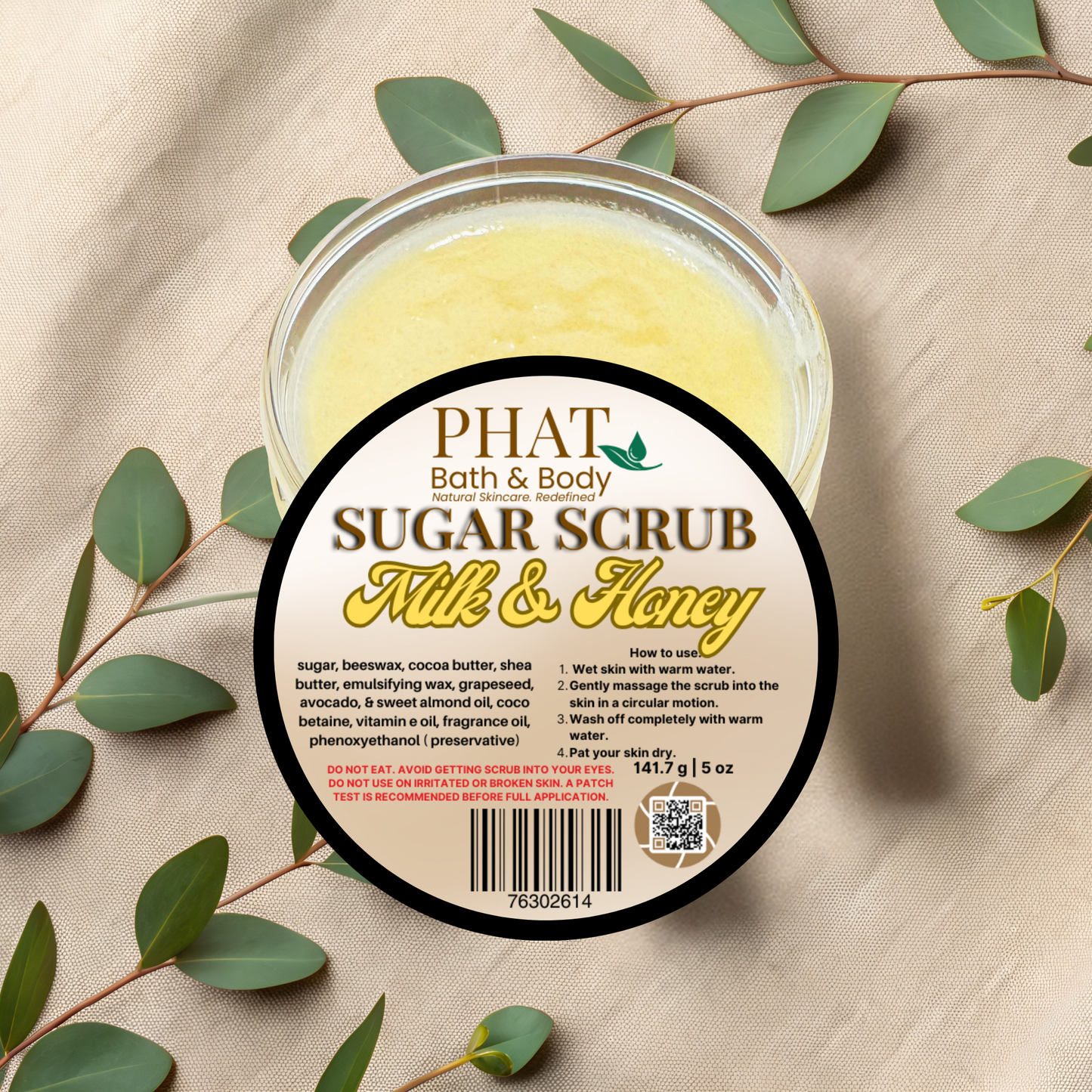 Jar of Phat Bath & Body Sugar Scrub with a label on a beige fabric background with green leaves.