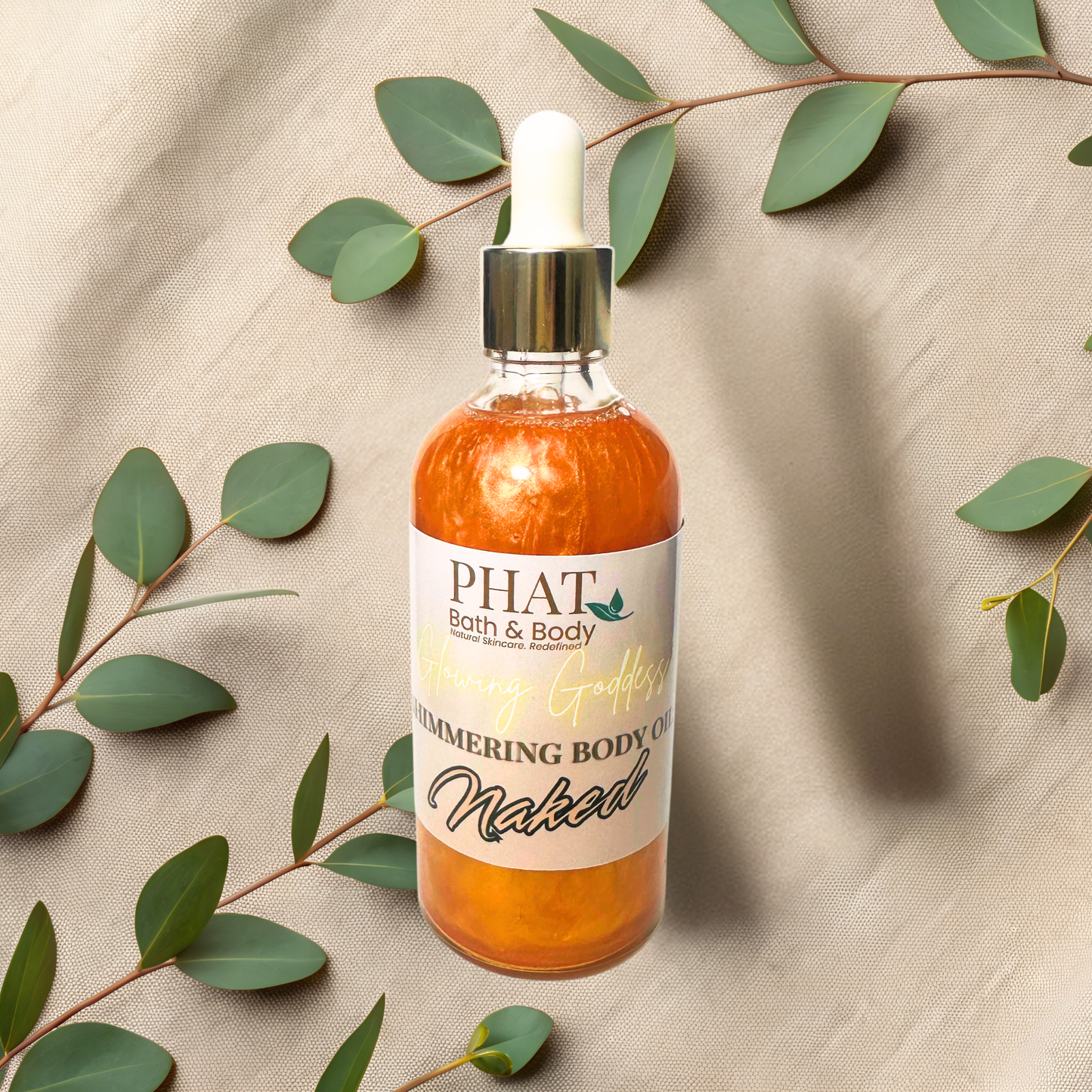 Bottle of 'Naked' simmering body oil from PHAT Bath & Body on a beige fabric background with green leaves.