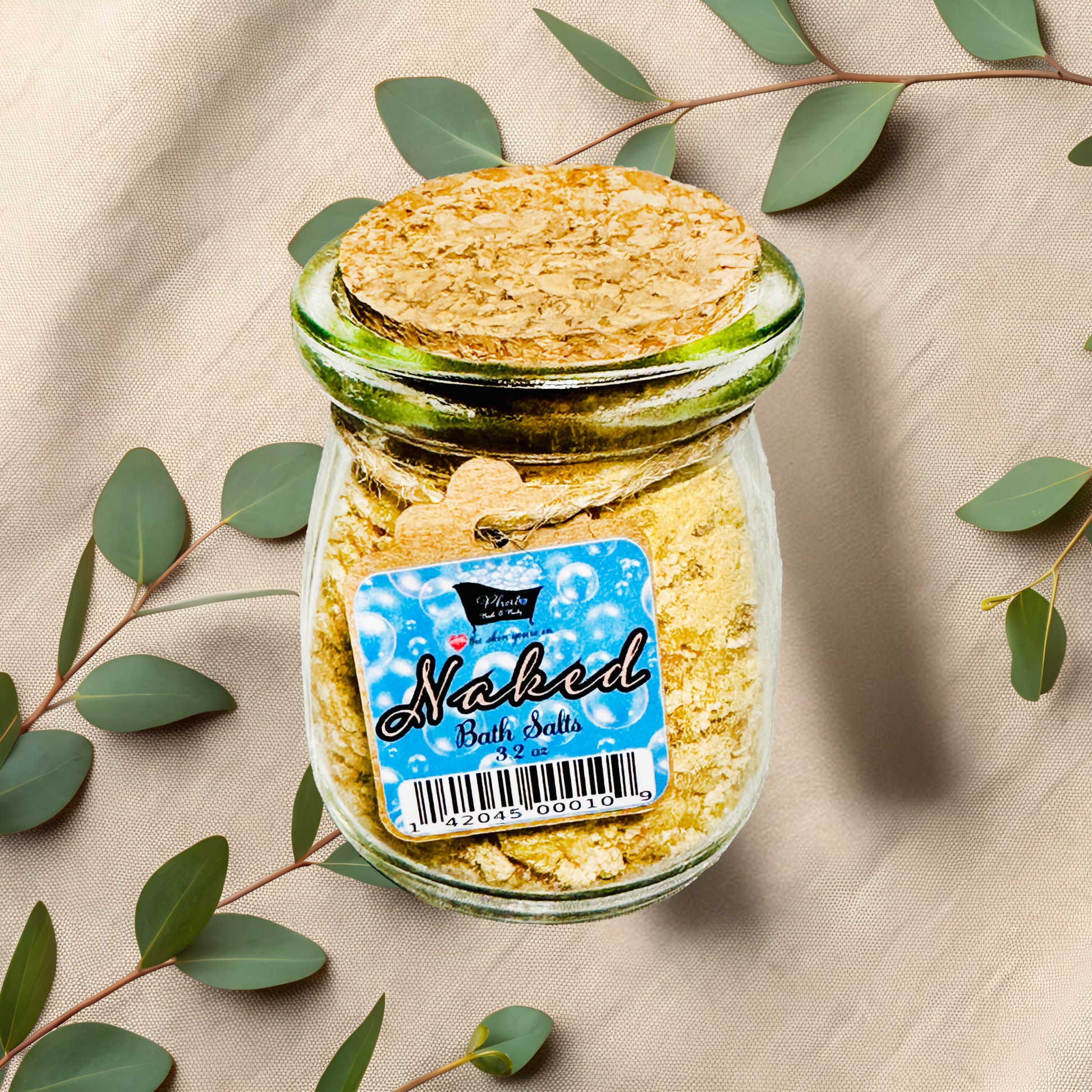 Jar of bath salts labeled 'Naked' with a cork lid on a beige fabric background with green leaves.