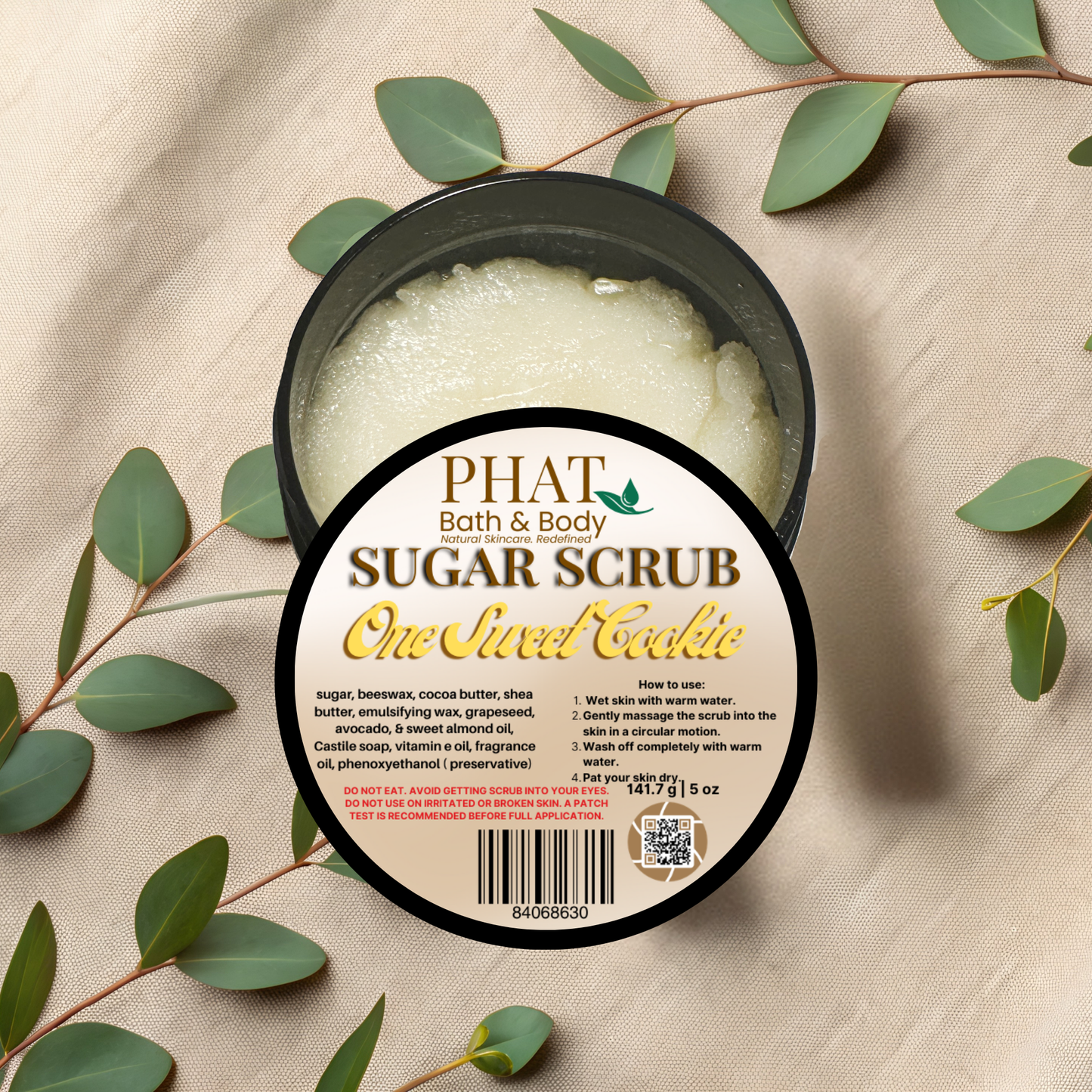 Jar of sugar scrub with a label on a beige fabric background with green leaves.