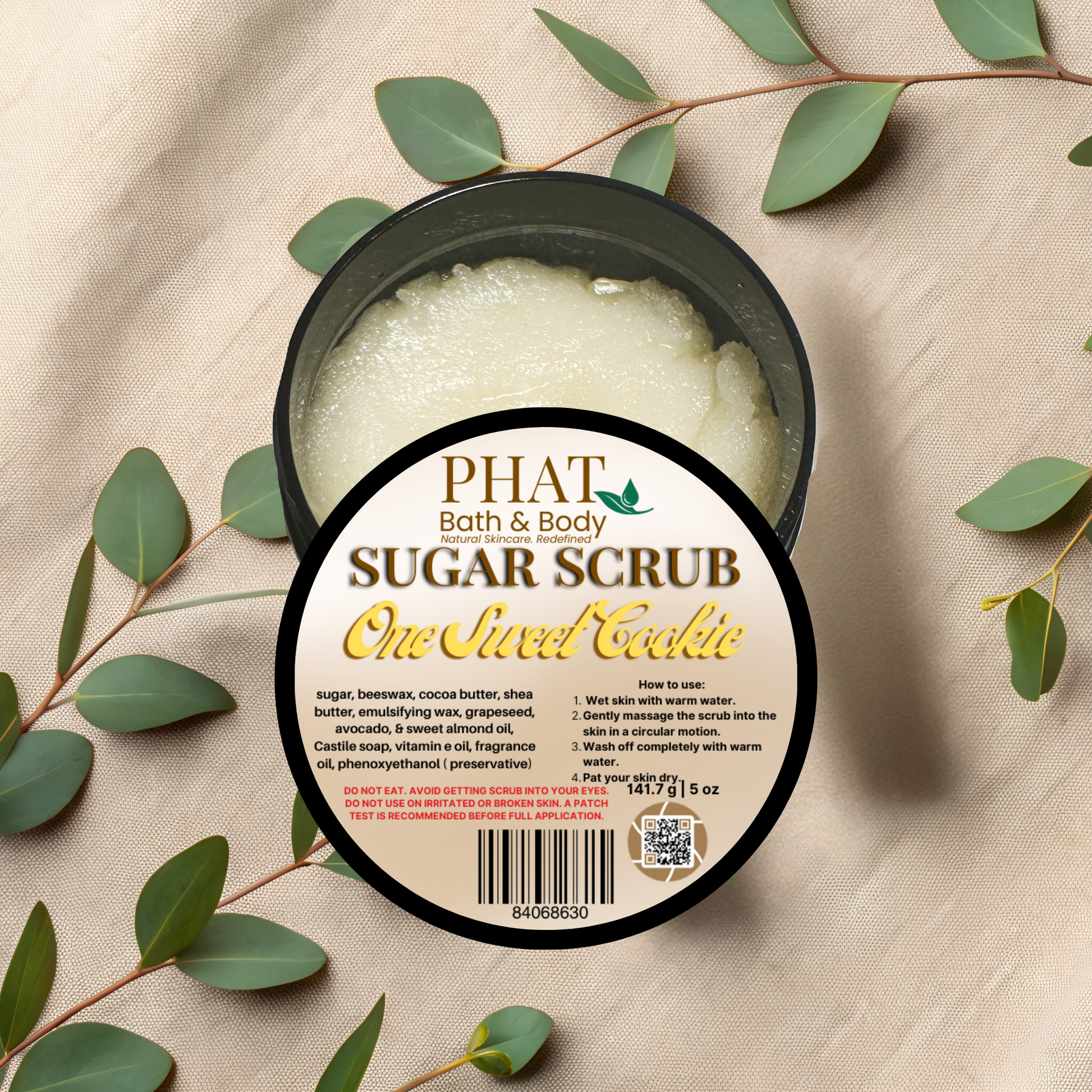 Jar of sugar scrub with a label on a beige fabric background with green leaves.
