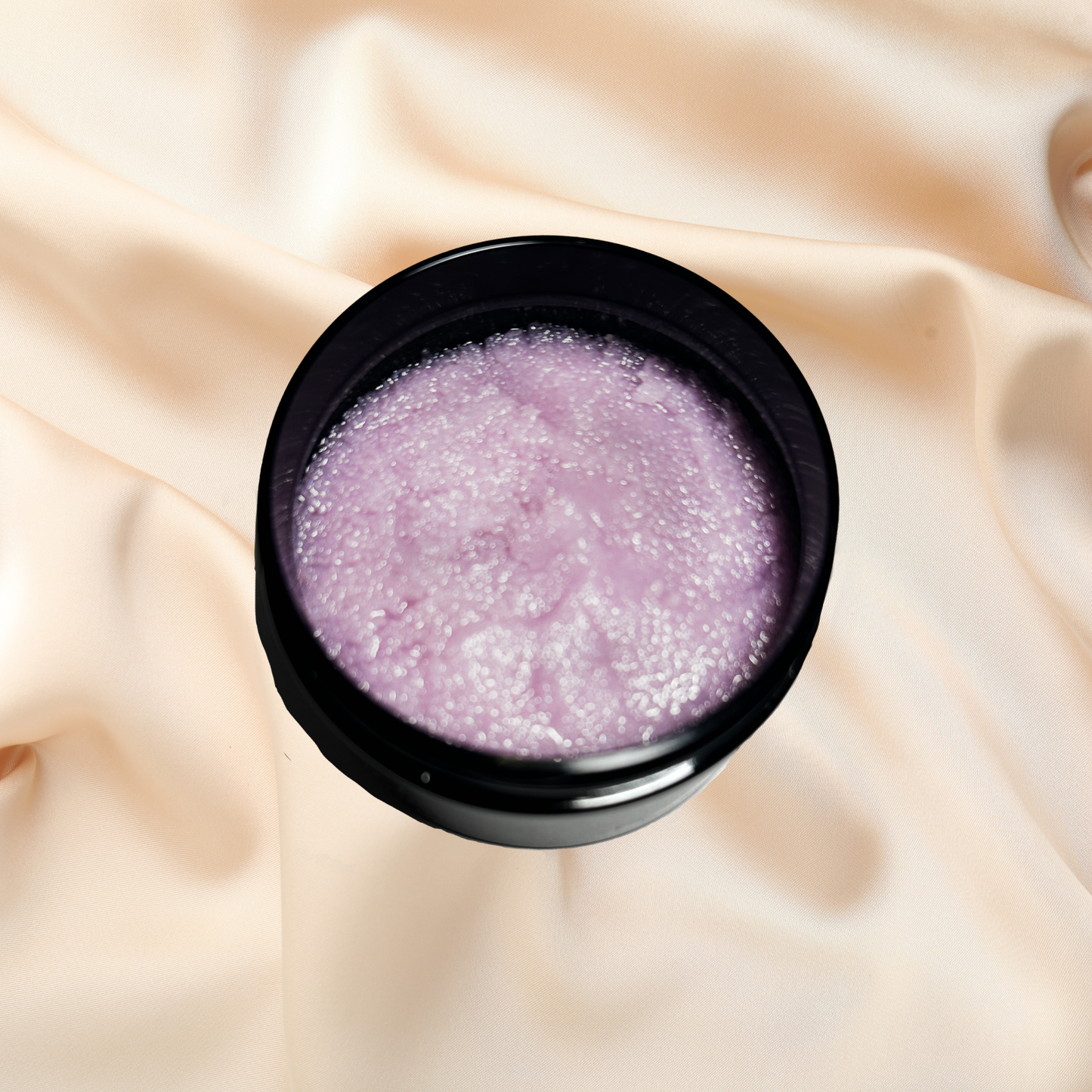 Black container with purple sugar scrub on a beige fabric background