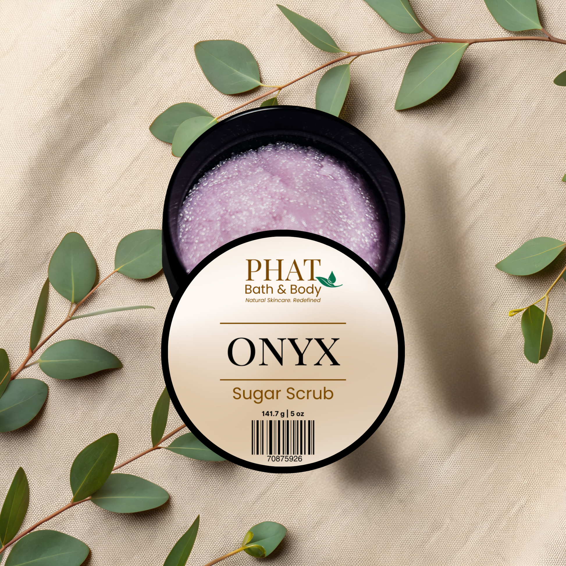 Body scrub jar labeled 'Onyx' by PHAT Bath & Body on a beige fabric background with green leaves.