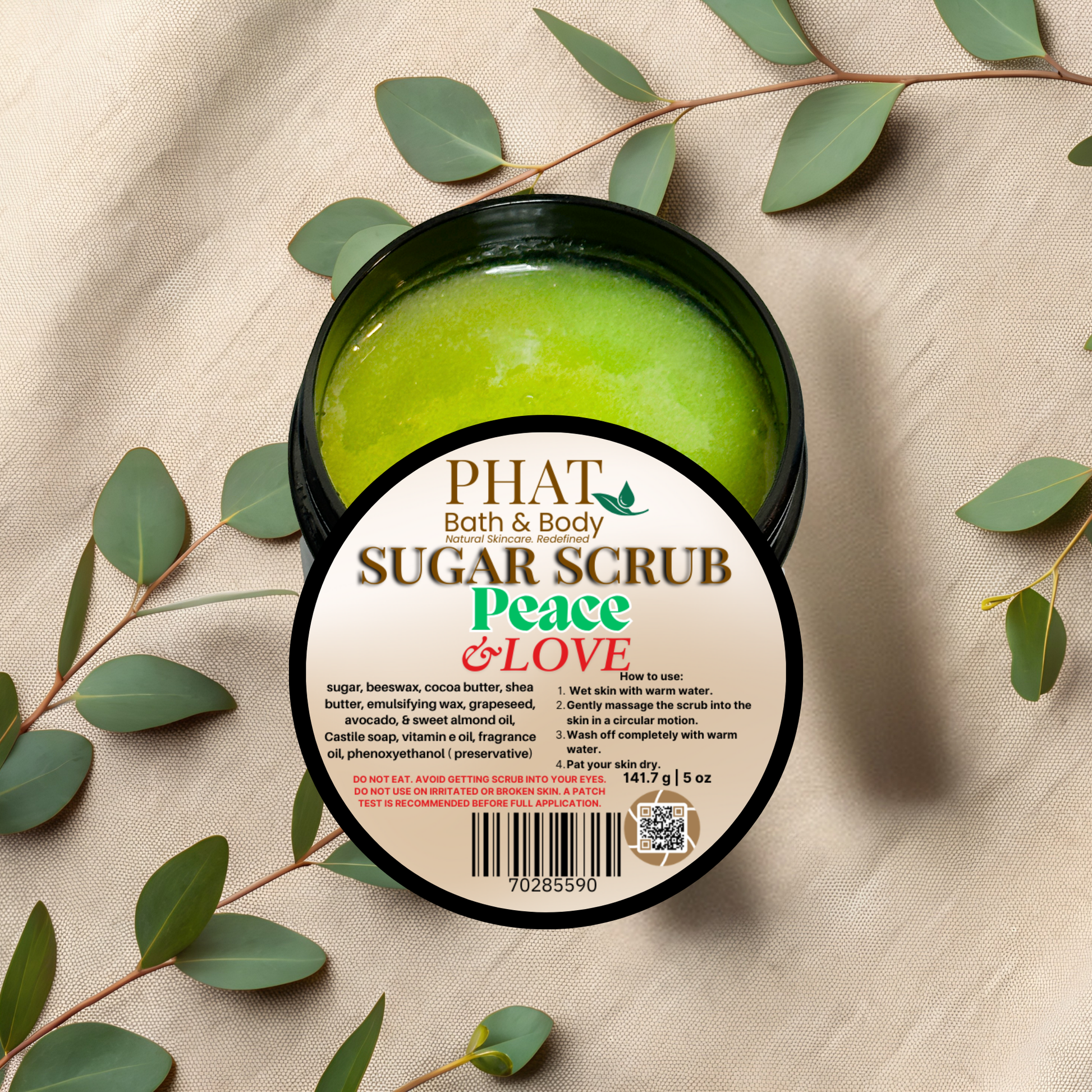 Phat Bath & Body sugar scrub container with green scrub inside, surrounded by eucalyptus leaves on a beige fabric background.