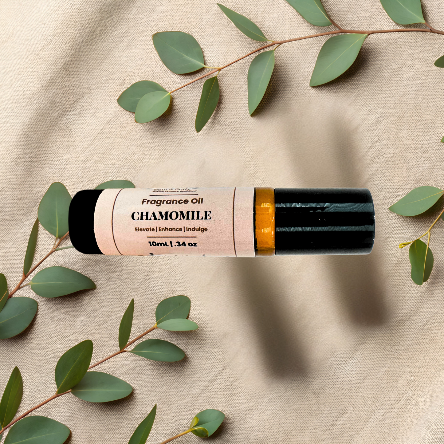 A 10ml roll-on bottle of Chamomile fragrance oil, styled on a beige textured fabric background with green eucalyptus leaves. The bottle has a black cap, a gold collar, and a cream-colored label with black text.