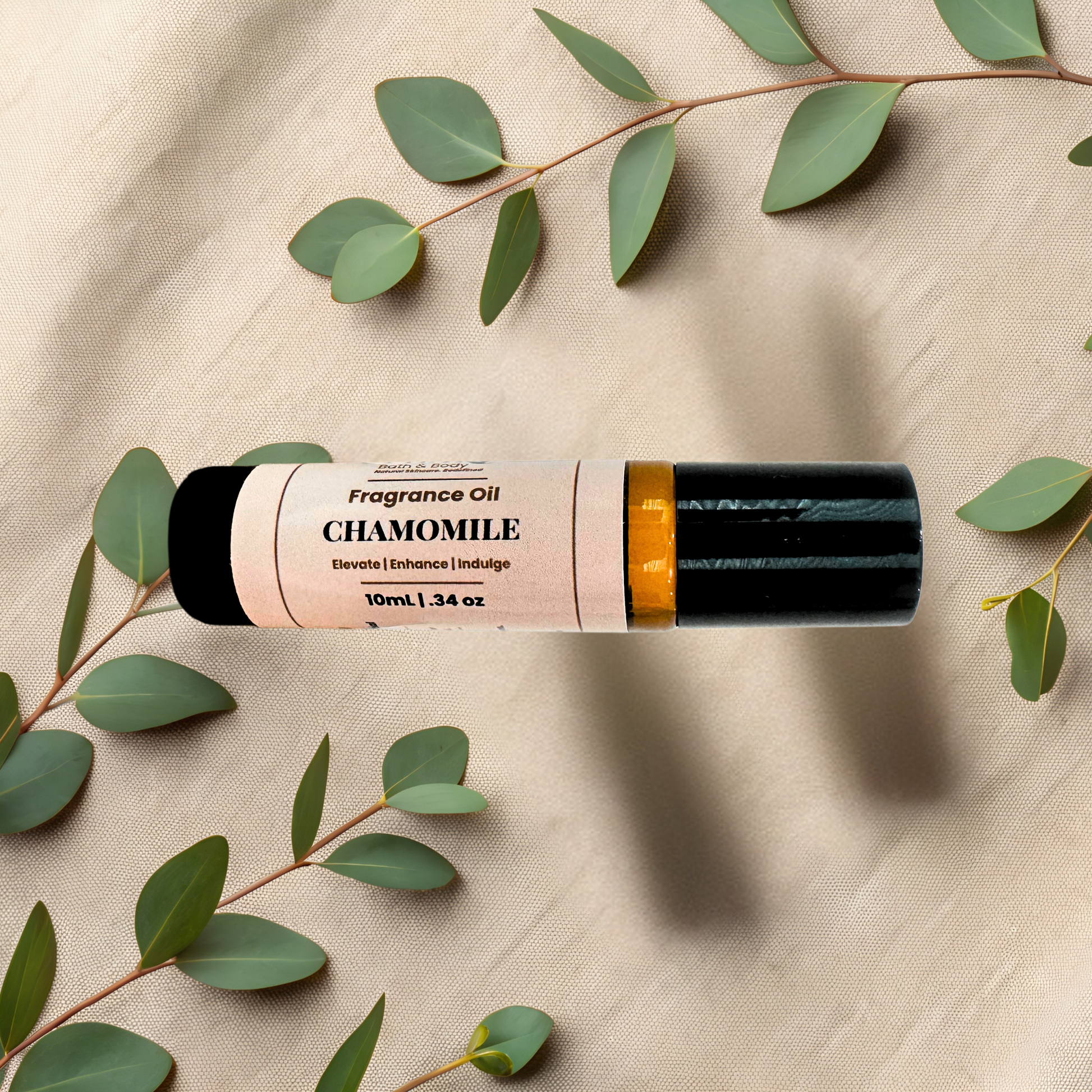 A 10ml roll-on bottle of Chamomile fragrance oil, styled on a beige textured fabric background with green eucalyptus leaves. The bottle has a black cap, a gold collar, and a cream-colored label with black text.