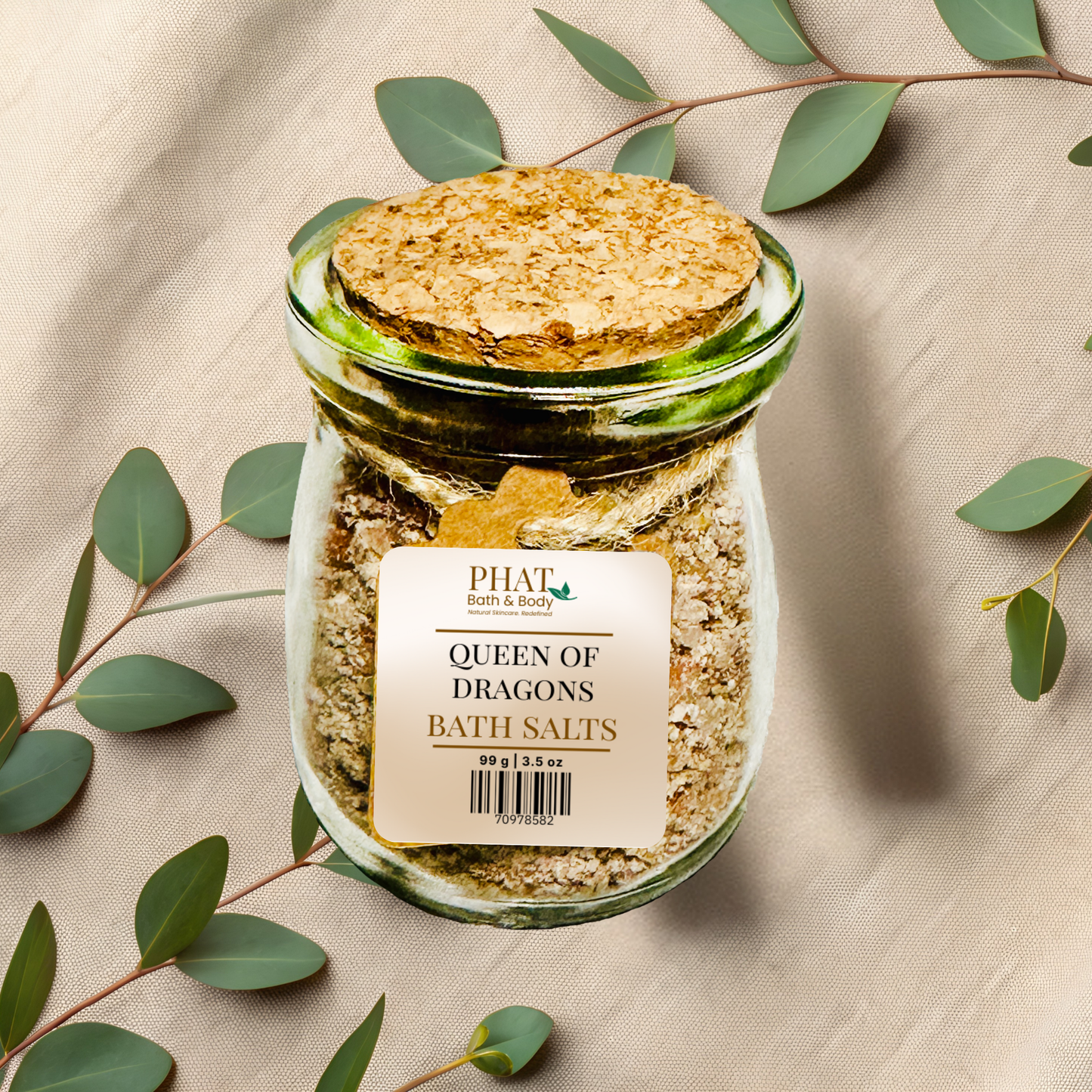 Jar of queen of dragons bath salts with a cork lid on a beige fabric background with green leaves.