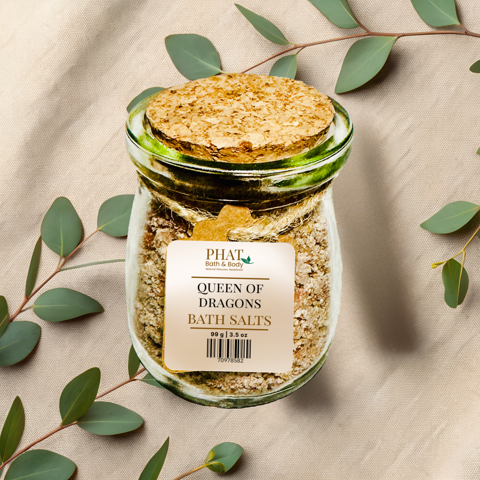 Jar of queen of dragons bath salts with a cork lid on a beige fabric background with green leaves.