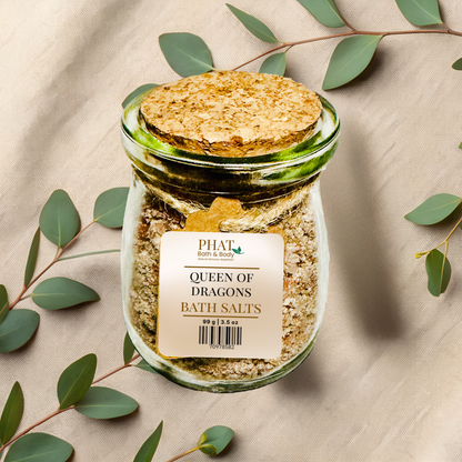 Jar of queen of dragons bath salts with a cork lid on a beige fabric background with green leaves.