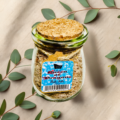 Jar of 'Queen of Dragons' bath salts with a cork lid on a beige fabric background with eucalyptus leaves.