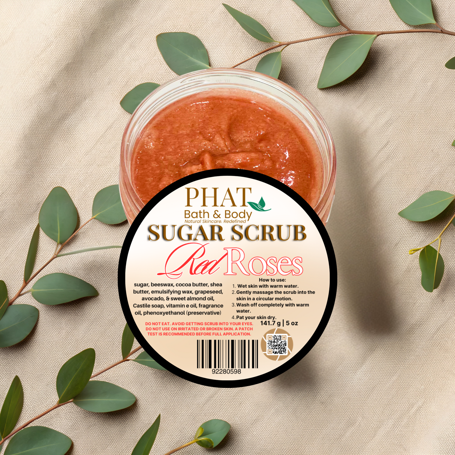 Jar of sugar scrub with 'Phat Bath & Body' label on a beige fabric background with green leaves.