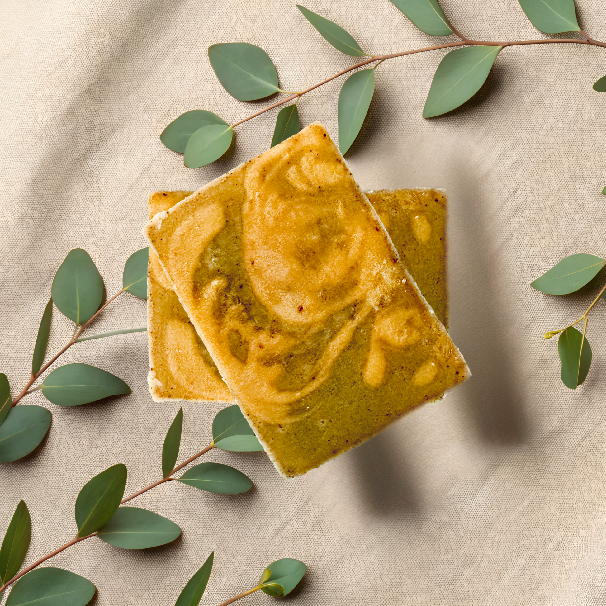 Two square yellowish-green bars of soap on a beige fabric background with green leaves.