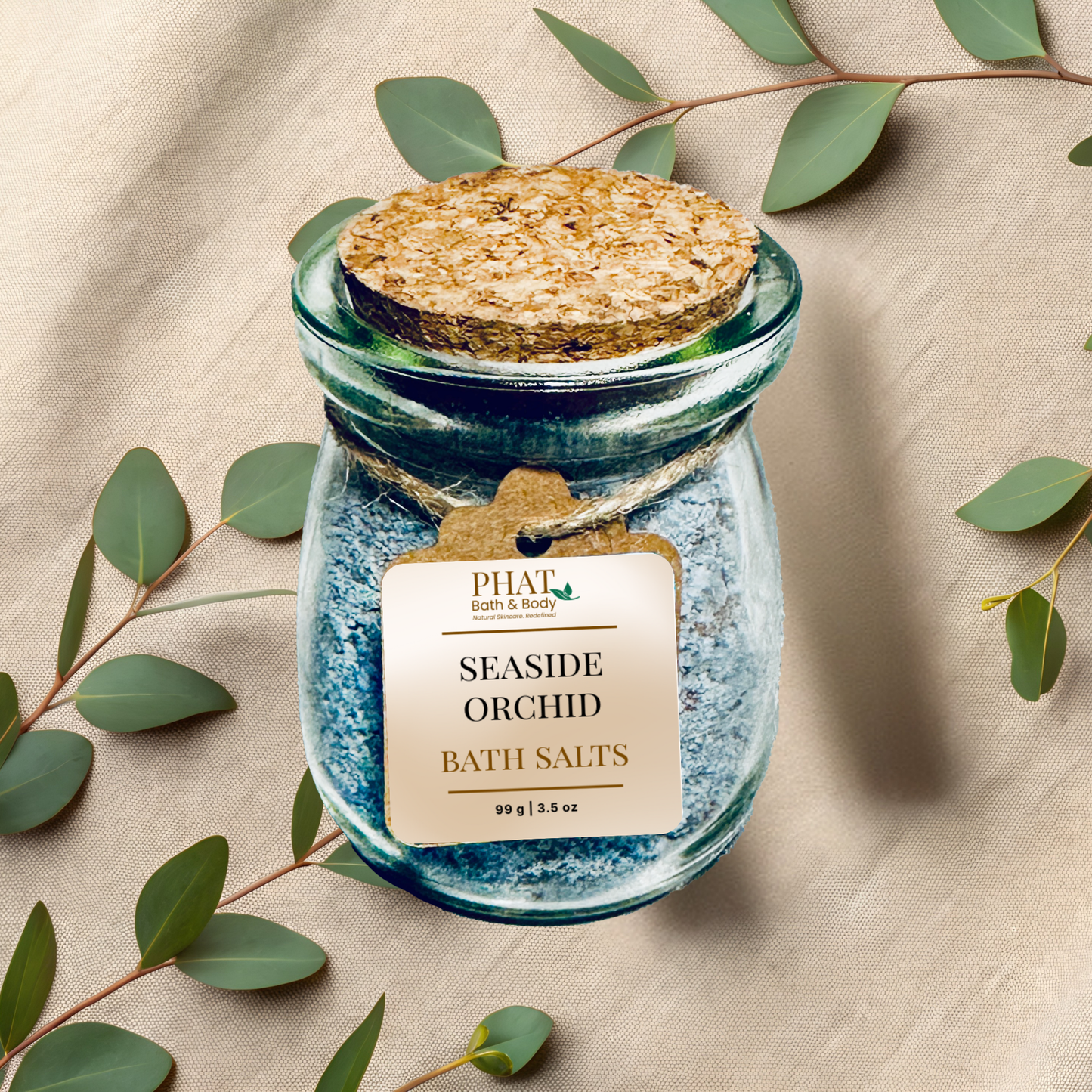 Jar of 'seaside orchid' bath salts with a cork lid on a beige fabric background with eucalyptus leaves.