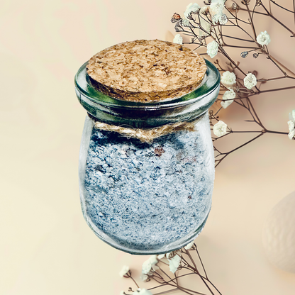 Glass jar with cork lid and blue bath salt, surrounded by dried flowers on a beige background