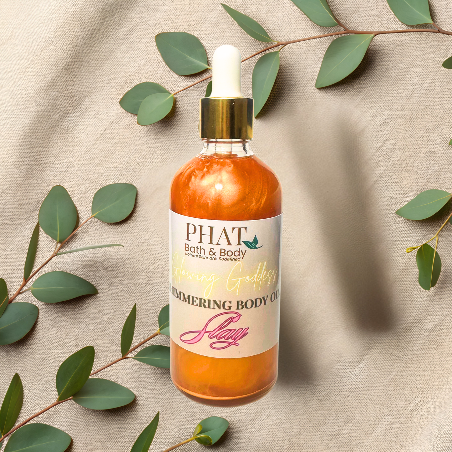 Bottle of 'PHAT Bath & Body' shimmering body oil with green leaves on a beige fabric background