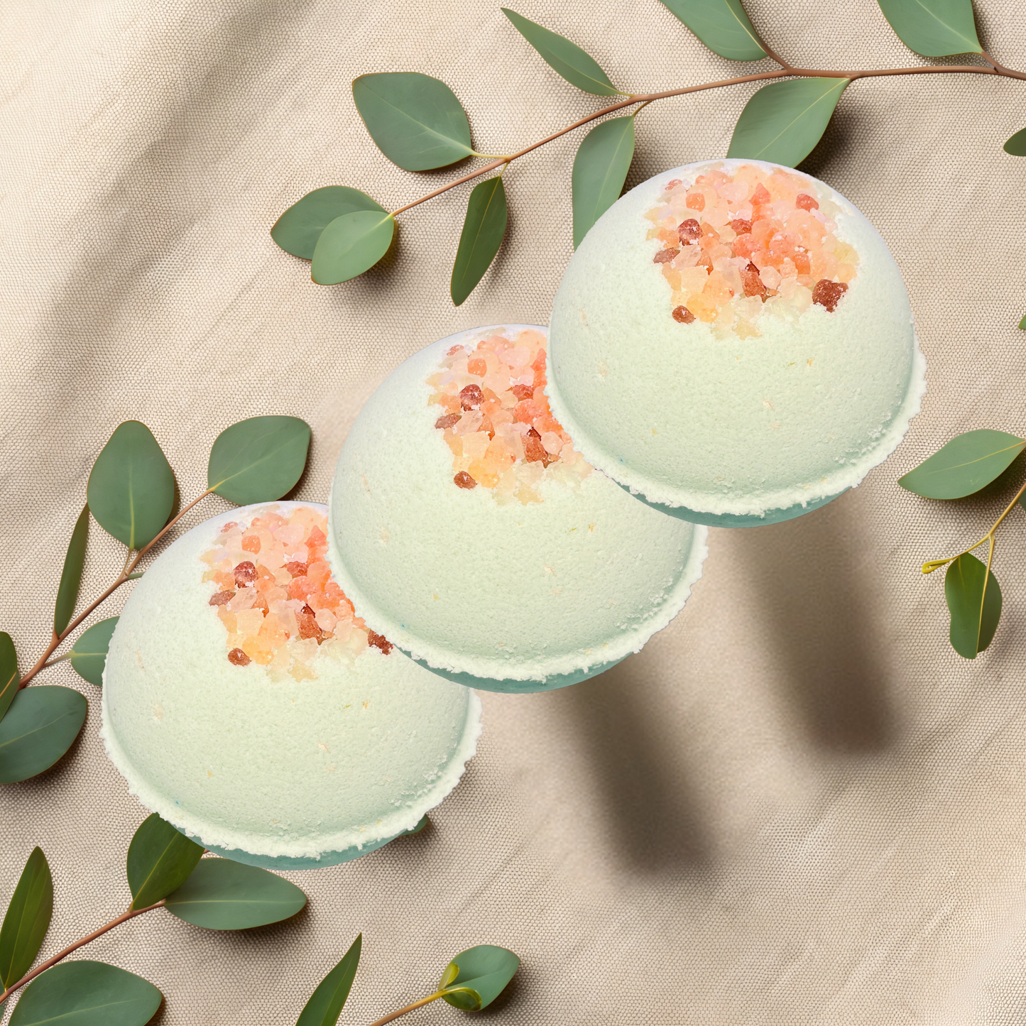 Three green bath bombs with pink salt on a beige fabric background with eucalyptus leaves.