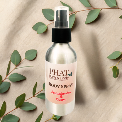 Body spray bottle labeled 'PHAT Bath & Body' with 'Strawberries & Cream' scent on a beige fabric background with green leaves.