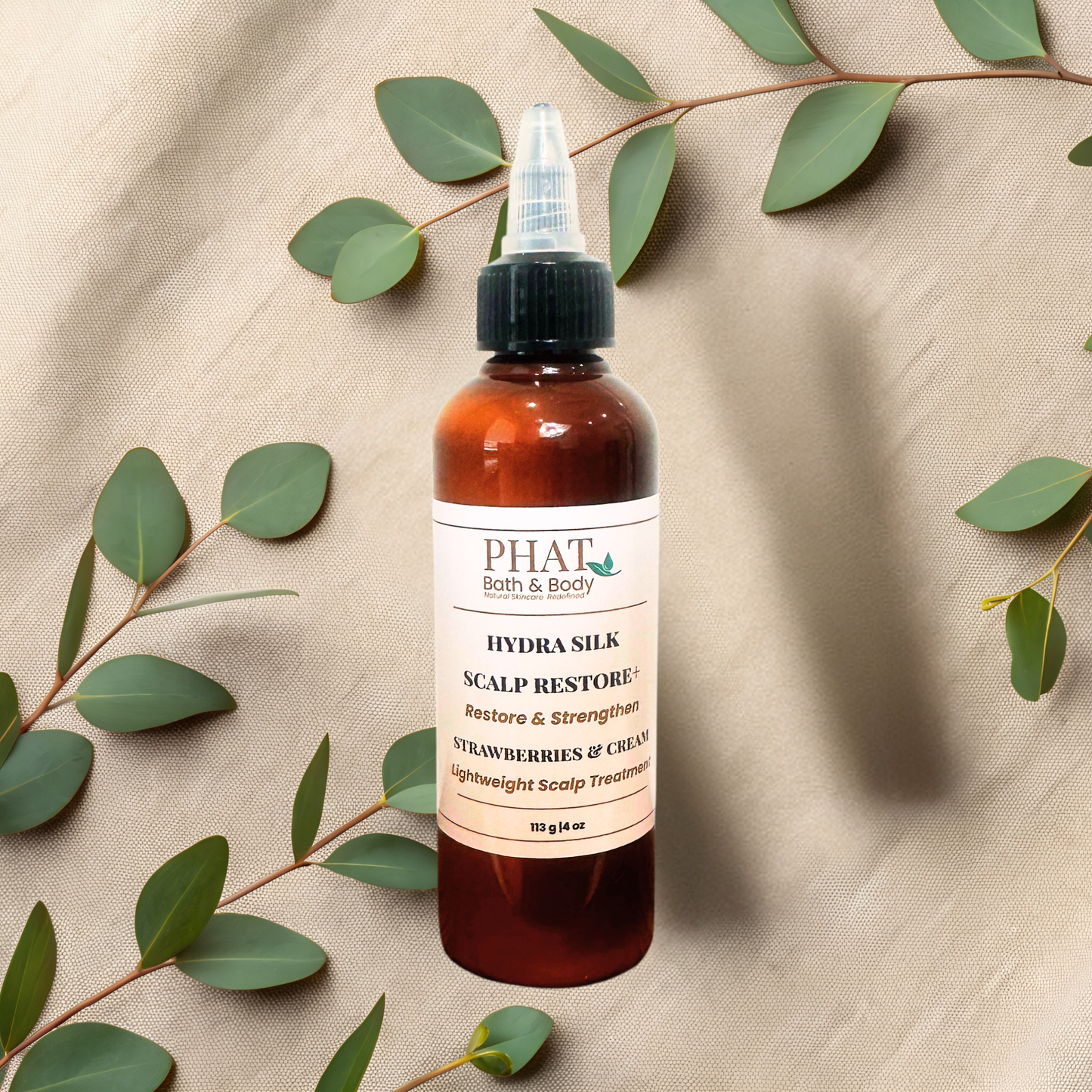 A bottle of PHAT Bath & Body Hydra Silk Scalp Restore+ scalp treatment, styled on a beige fabric background with eucalyptus leaves. The bottle is amber brown with a cream label and a black cap.