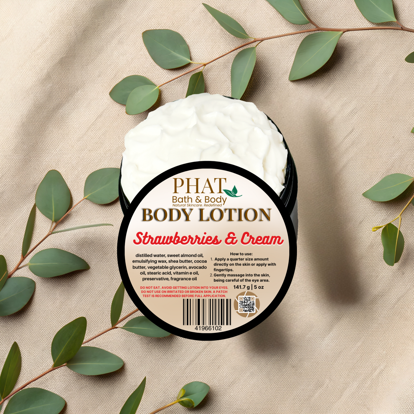 Body lotion container labeled 'Strawberries & Cream' by Phat Bath & Body on a beige fabric background with green leaves.