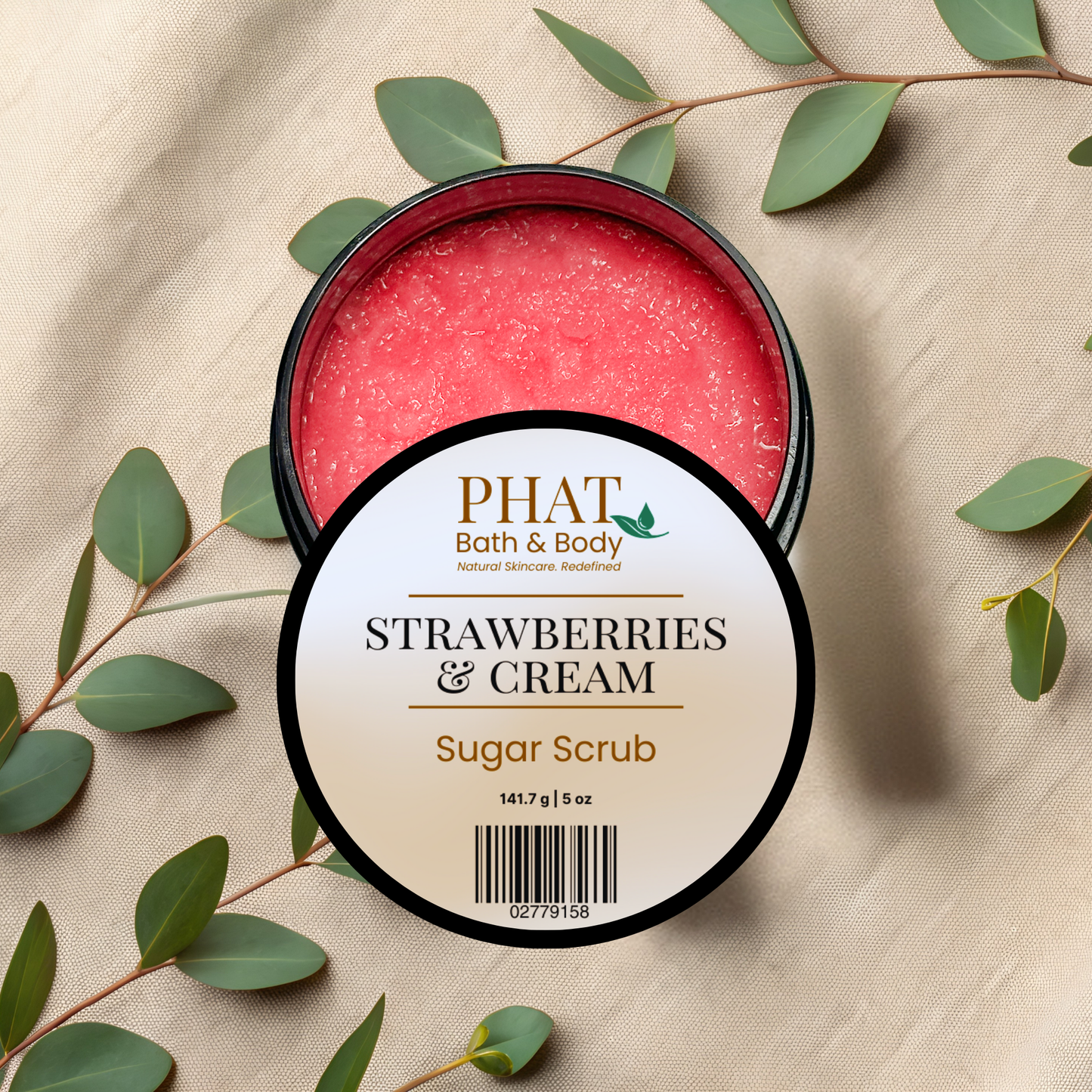 An open round black tub of PHAT Bath & Body Strawberry & Cream Sugar Scrub, showing the pink-red scrub inside, with a round white label in front. The tub is styled on a beige fabric background with green eucalyptus leaves.