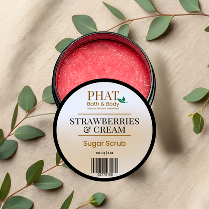 An open round black tub of PHAT Bath & Body Strawberry & Cream Sugar Scrub, showing the pink-red scrub inside, with a round white label in front. The tub is styled on a beige fabric background with green eucalyptus leaves.