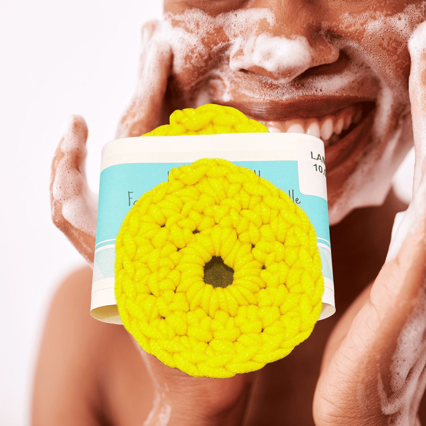 Yellow round scrubby with a brand label held by a person with soap suds on their face.