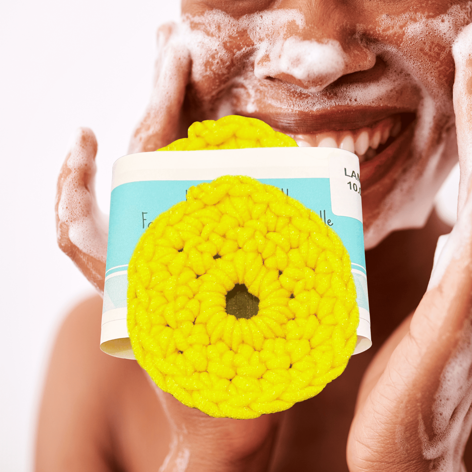 Yellow round scrubby with a brand label held by a person with soap suds on their face.