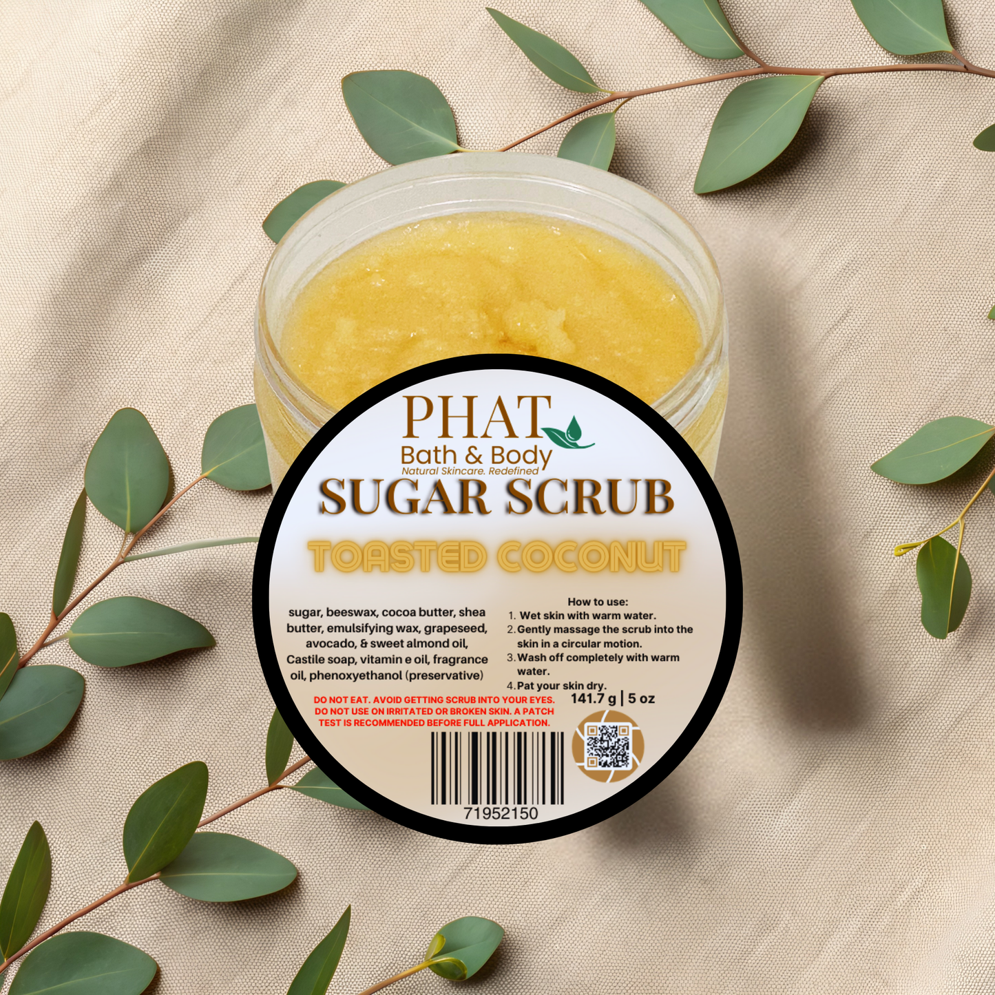 Jar of Phat Bath & Body Sugar Scrub with toasted coconut flavor on a beige fabric background with green leaves.