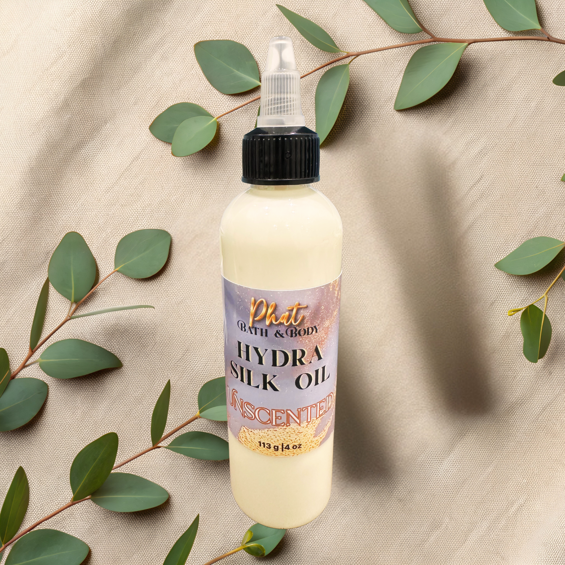 Bottle of Phat Bath & Body Hydra Silk Oil with green leaves on a beige fabric background