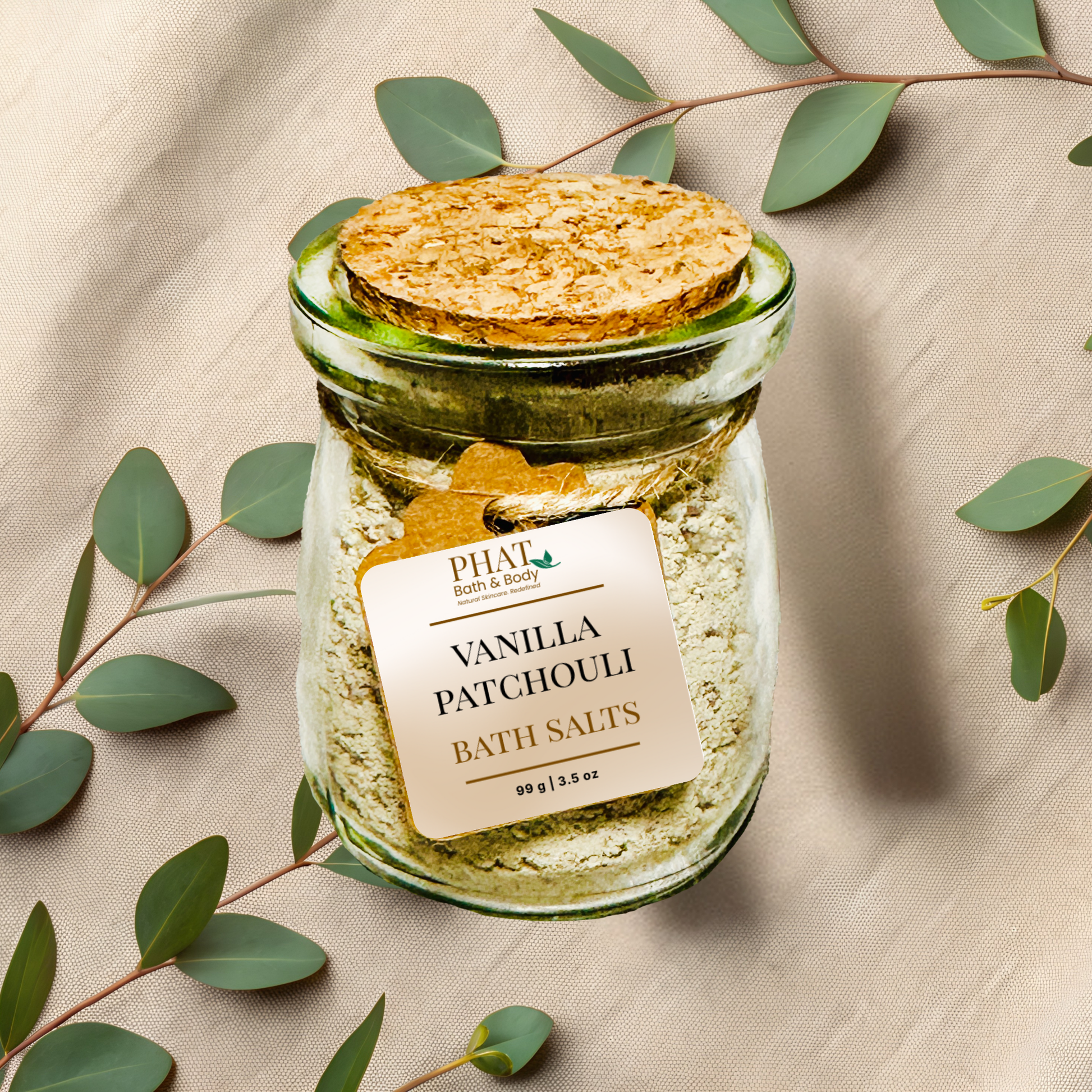 Jar of 'vanilla patchouli' bath salts with a cork lid on a beige fabric background with eucalyptus leaves.