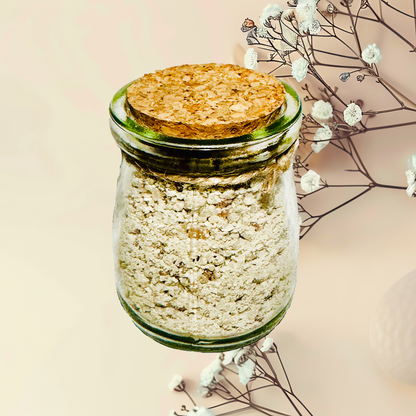Glass jar with cork lid containing a light-colored bath salts, surrounded by delicate white flowers on a soft pink background.