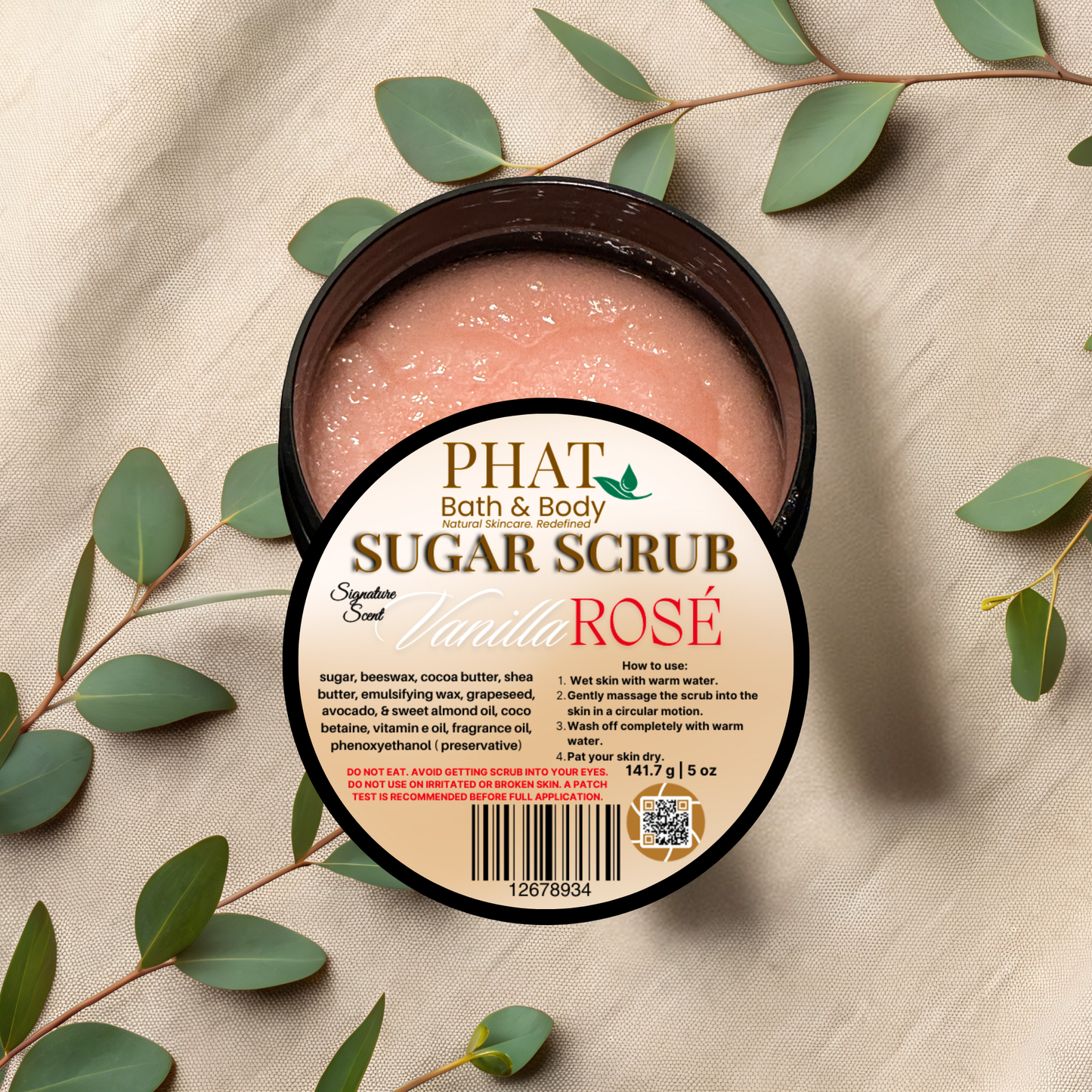 Jar of Phat Bath & Body Sugar Scrub with a label on a beige fabric background with green leaves.