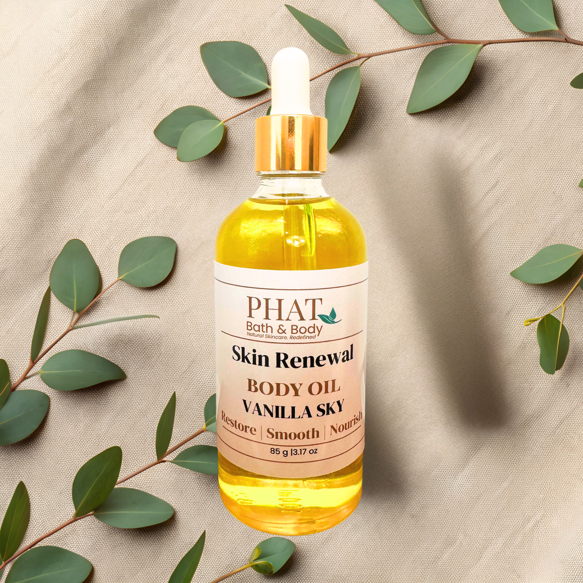 A bottle of PHAT Bath & Body Skin Renewal Body Oil in Vanilla Sky scent, filled with golden yellow oil and featuring a white dropper cap with a gold collar. The bottle is centered on a beige textured fabric background decorated with green eucalyptus leaves.