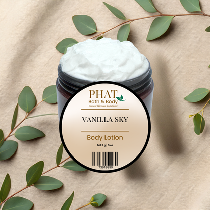 A jar of PHAT Bath & Body Vanilla Sky Body Lotion with white whipped lotion visible at the top, set on a beige fabric background with eucalyptus leaves around it.