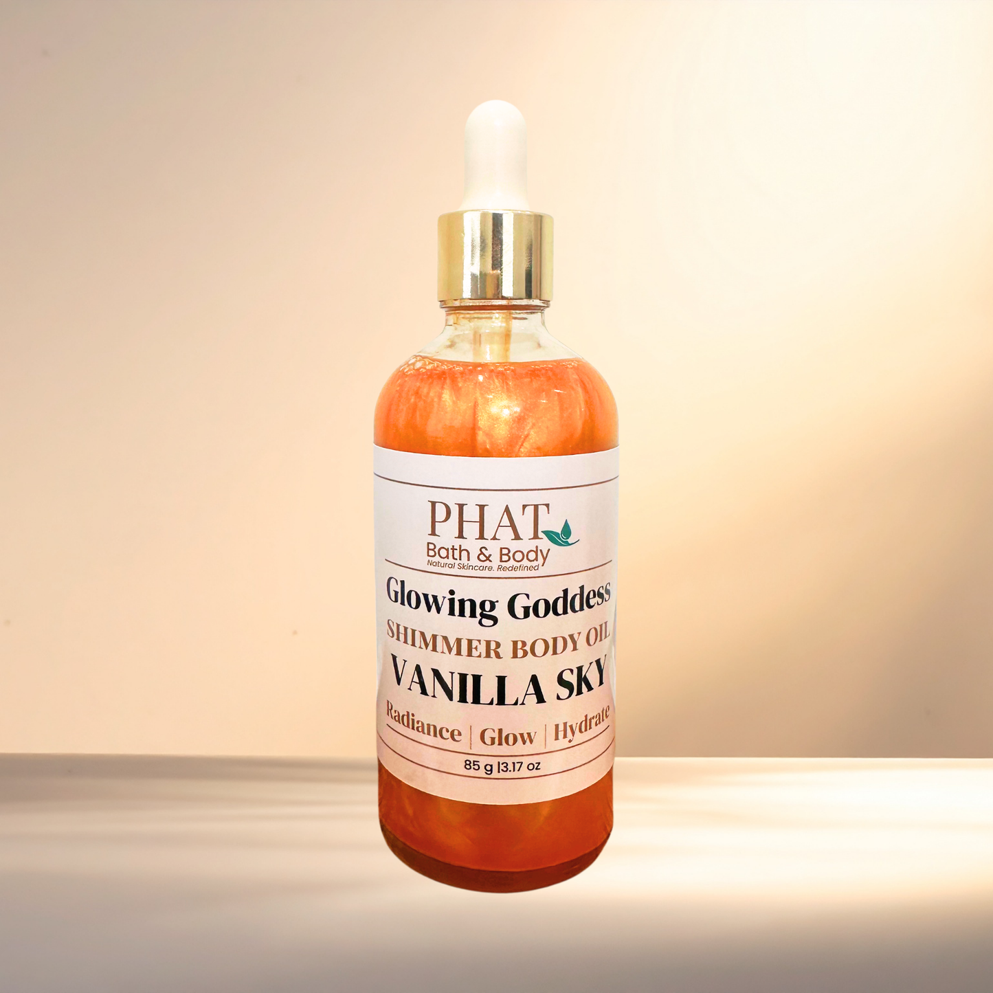 A bottle of PHAT Bath & Body 'Glowing Goddess' shimmer body oil in Vanilla Sky, featuring an amber-colored liquid, a white label, and a gold dropper cap, set against a warm beige background.