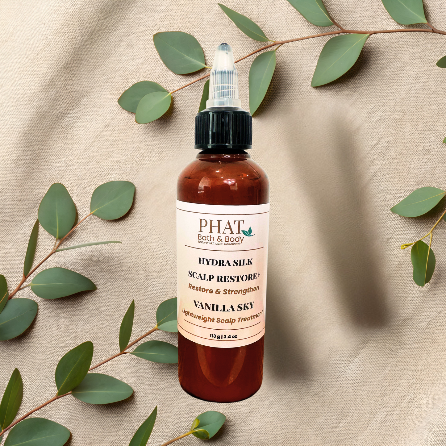 A bottle of PHAT Bath & Body Hydra Silk Scalp Restore + Restore & Strengthens in Vanilla Sky scent, styled on a beige fabric background with eucalyptus leaves.