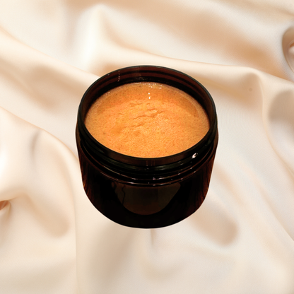 An open dark amber cosmetic jar filled with a smooth, brown scrub, set against a draped cream satin fabric background.