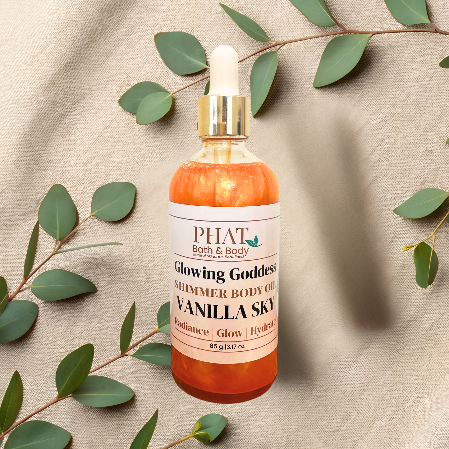 A bottle of PHAT Bath & Body 'Glowing Goddess' shimmer body oil, Vanilla Sky, filled with orange liquid and topped with a white dropper cap and gold collar, styled on a beige fabric background with green eucalyptus leaves.