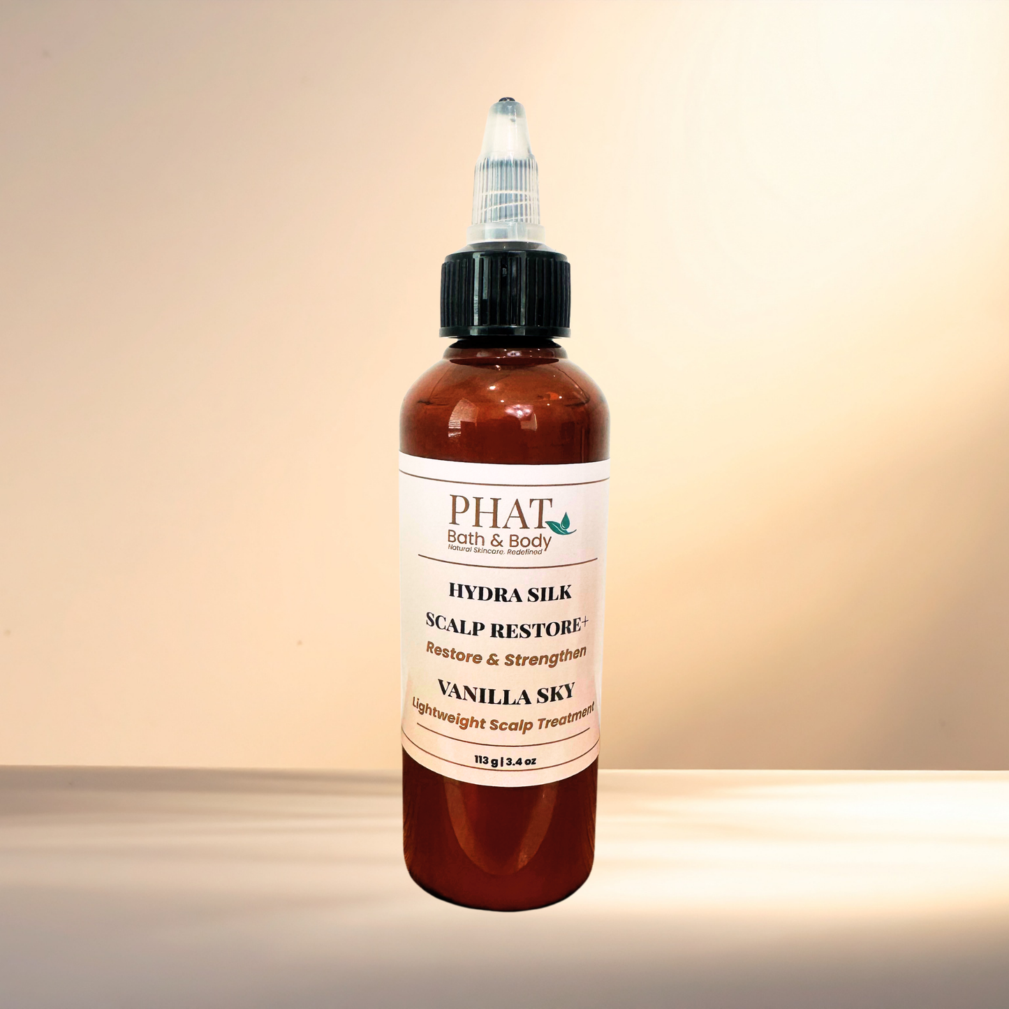 A bottle of PHAT Bath & Body Hydra Silk Scalp Restore + in Vanilla Sky scent, featuring an amber brown bottle, black dropper cap, and a white label, set against a warm beige background.
