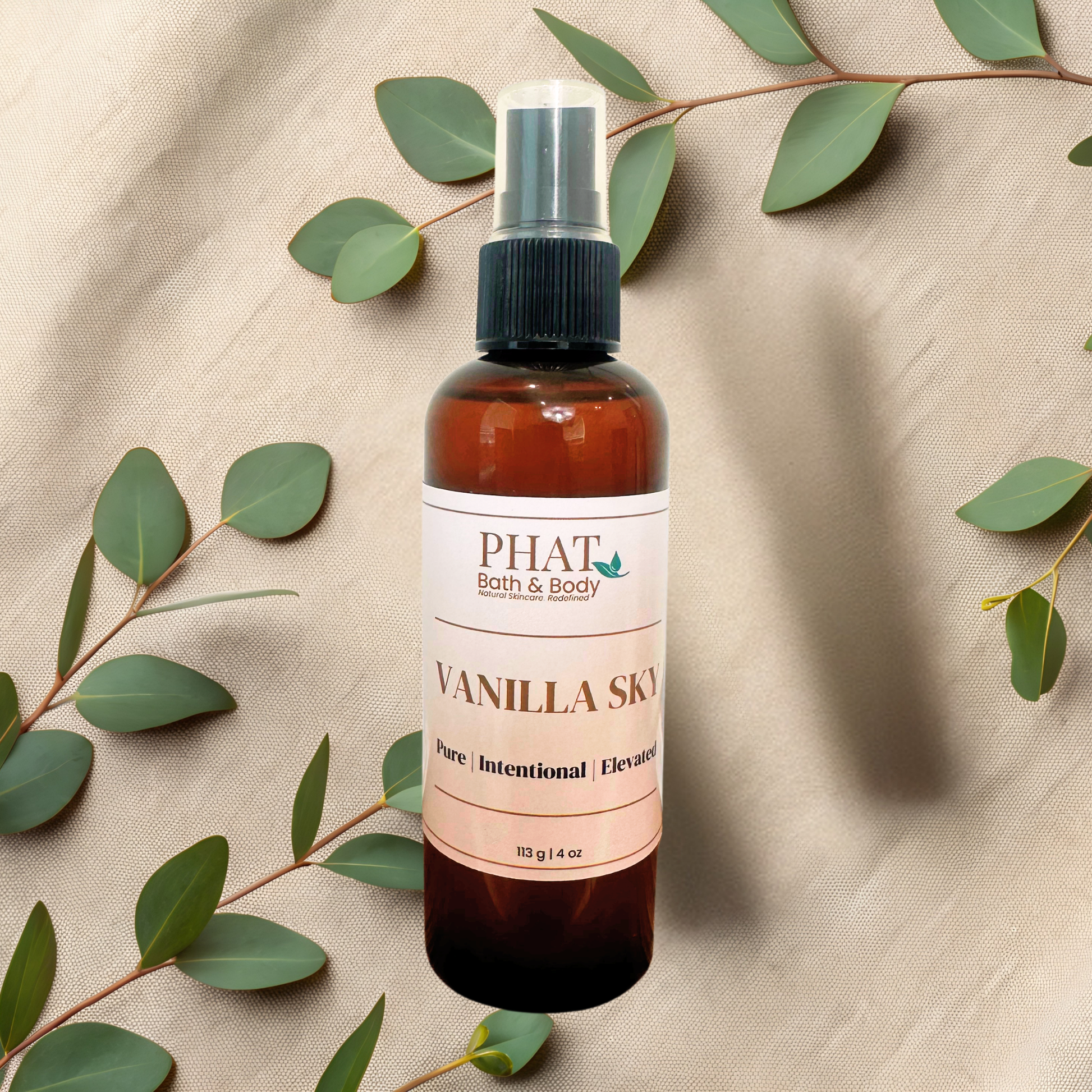 An amber spray bottle of PHAT Bath & Body Vanilla Sky body mist, styled on a beige fabric background with eucalyptus leaves.