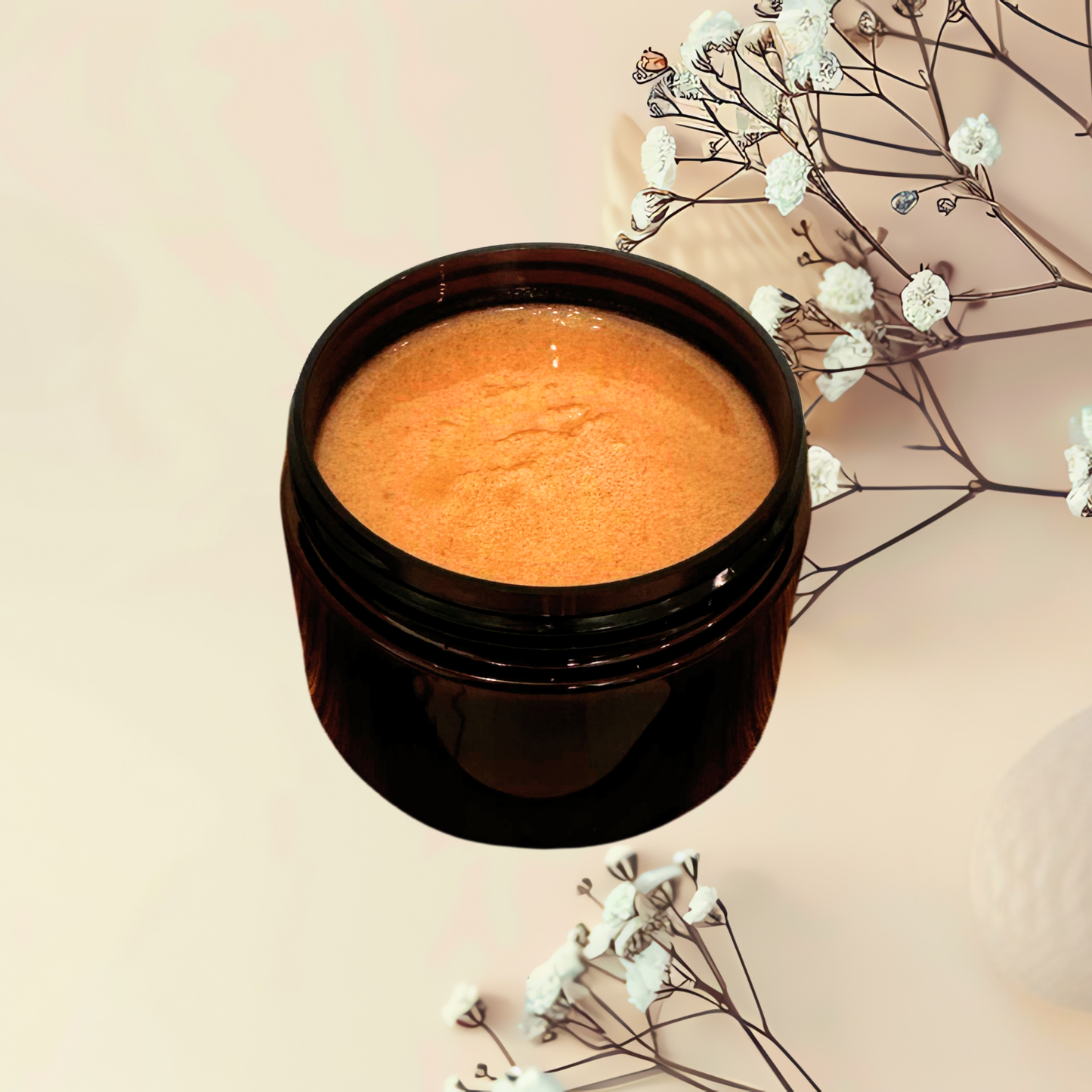 An open dark amber cosmetic jar filled with a textured brown scrub, styled on a warm beige background with delicate white flowers.