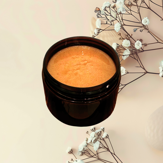 An open dark amber cosmetic jar filled with a textured brown scrub, styled on a warm beige background with delicate white flowers.