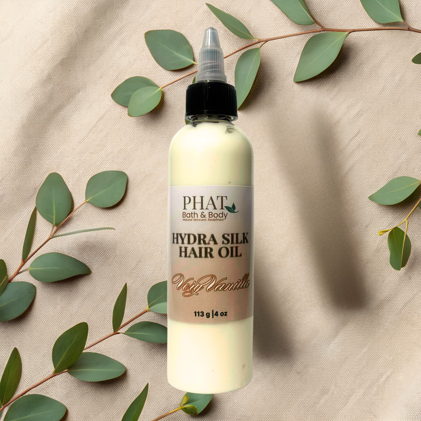 Bottle of Phat Bath & Body Hydra Silk Hair Oil on a beige fabric background with green leaves.