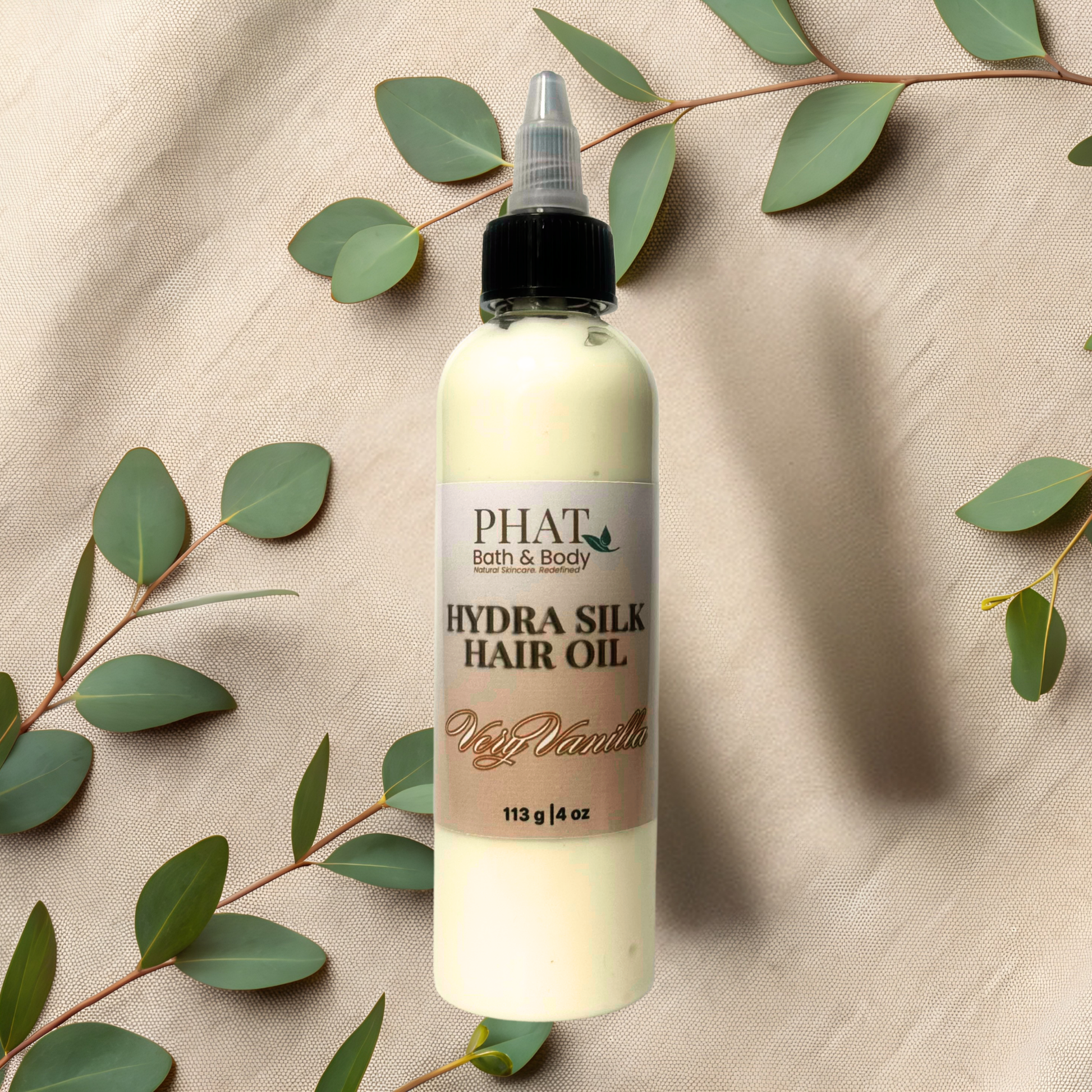 Bottle of Phat Bath & Body Hydra Silk Hair Oil on a beige fabric background with green leaves.