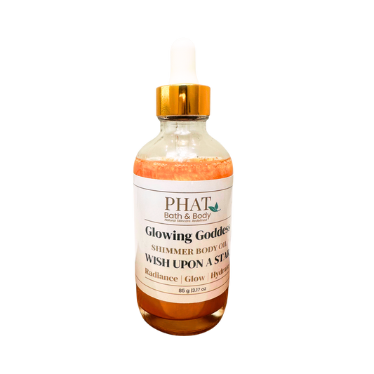 A clear glass bottle of PHAT Bath & Body 'Glowing Goddess' shimmer body oil with amber liquid, a white label, and a gold dropper cap, isolated on a white background.